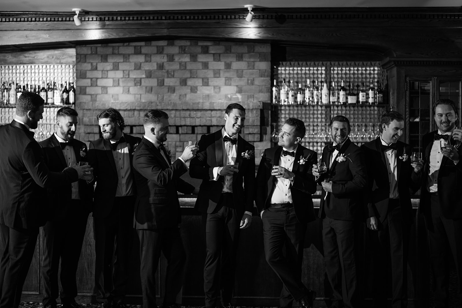Black and white film photo of the groom and groomsmen sharing a laugh and toasting at the bar.